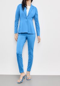 Vaaleansininen puku, jossa on muotonsa mukainen bleiseri, jossa on hapsuolkaimet, sekä siihen sopivat kapealahkeiset housut. Samettimainen pinta, jota täydentävät siniset strappy-korkokengät.