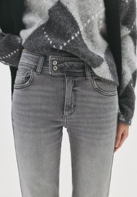 Osoba oblečená v svetlosivých rifliach s vysokým pásom a dvojitým zapínaním, skombinovaných s huňatým čiernym a sivým svetrom s vzorom.