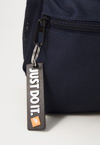 Tamsiai mėlynas audinio krepšys su juodu užtrauktuku, ant kurio yra juoda etiketė su užrašu „JUST DO IT.“ ir mažu oranžiniu Nike logotipu.