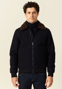 Ciemnoniebieska kurtka z brązowym kołnierzem z owczej wełny, zapinana na zamek błyskawiczny, z ściągaczami przy mankietach. Materiał jest gładki o lekkiej strukturze.