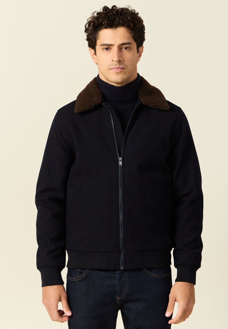 Ciemnoniebieska kurtka z brązowym kołnierzem z owczej wełny, zapinana na zamek błyskawiczny, z ściągaczami przy mankietach. Materiał jest gładki o lekkiej strukturze.