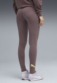 Person, der mauve Leggings und lilafarbene Puma-Sneaker mit weißen Socken trägt, steht vor einem einfarbigen grauen Hintergrund.