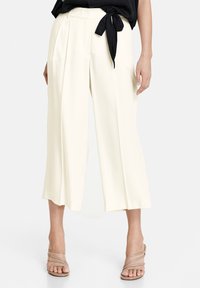 Wijde broek in een licht crèmekleurige stof, voorzien van plooien en een zwarte stoffen riem, gecombineerd met beige sandalen met straps.