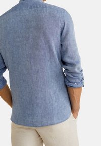 Hellblaues Leinenhemd mit strukturierter Oberfläche, geknöpften Manschetten und klassischem Kragen. Der Rücken zeigt einen geraden Saum und minimalistische Details.