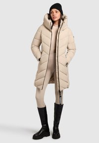 Beige gewatteerde jas met een hoge kraag en ritsluiting, voorzien van een chevron ontwerp. Gecombineerd met zwarte kniehoge laarzen en beige leggings.