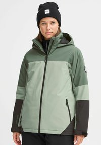 Grüne und schwarze wasserdichte Jacke mit Reißverschluss, Kapuze und zwei Seitentaschen. Sie hat eine glatte Textur und ein mehrschichtiges Farbdesign.