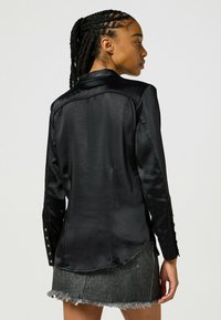 Czarna satynowa koszula z wykładanym kołnierzykiem, zapiętymi mankietami i zaokrąglonym dołem. Tkanina ma gładką strukturę i subtelny blask.