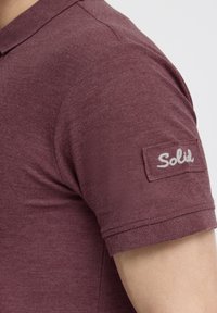 Bordeaux poloshirt i struktureret stof, med en rund krave og et mærke på ærmet med "Solid" broderet i hvid.