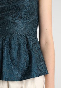 Haut en dentelle bleu sarcelle avec un ourlet à basque, présentant des motifs floraux complexes et un tissu texturé. Le design est structuré et ajusté à la taille.