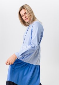 Lichtblauw overhemdjurk met een gestreepte bovensectie en een vloeiende blauwe rok. Lange mouwen met knoopmanchetten. Zachte stof structuur.