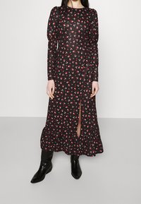 Kvinna i en svart långärmad midi-klänning med små röda och vita blommor, fram-slit, volangkant och svarta spetsiga stövlar.