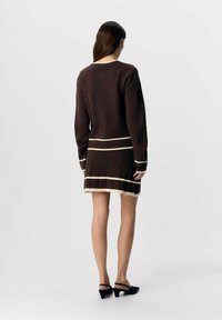 Brun stickad klänning med krämfärgade ränder, med ribbad textur och en veckad kjol. Modellen bär svarta slingback-klackar.