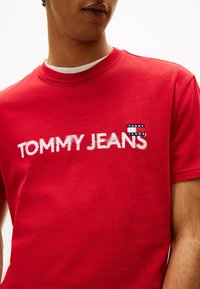 Rotes Baumwoll-T-Shirt mit dem "TOMMY JEANS" Logo in Weiß und Schwarz auf der Vorderseite. Kurze Ärmel und runder Halsausschnitt, glatte Textur.