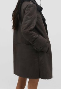 Cappotto in pelle marrone con una finitura testurizzata. Presenta un colletto a punta, polsini con bottoni e tasche laterali. Arriva fino a metà coscia.