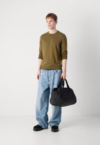 Sac de sport synthétique noir avec une forme arrondie, deux poignées et un petit logo rouge. Le modèle porte un pull vert olive et un jean baggy bleu clair.