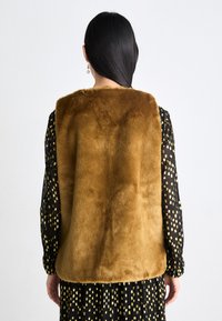 Braune Kunstfellweste mit weicher Textur, ausgestattet mit einem runden Ausschnitt und ärmellosem Design, getragen über einem schwarzen Kleid mit goldenen Mustern.