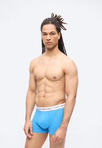 Άνδρας με dreadlocks που φοράει μπλε εσώρουχα Tommy Hilfiger στέκεται μπροστά σε λευκό φόντο, δείχνοντας έναν μυώδη κορμό.