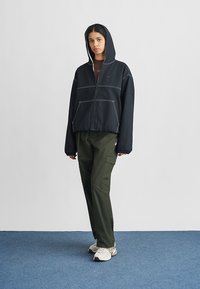 Μαύρο φούτερ με φερμουάρ και αντίθετες λευκές ραφές, με κουκούλα και πλαϊνές τσέπες, σε συνδυασμό με παντελόνι cargo σε ελαιί πράσινο και λευκά αθλητικά παπούτσια.