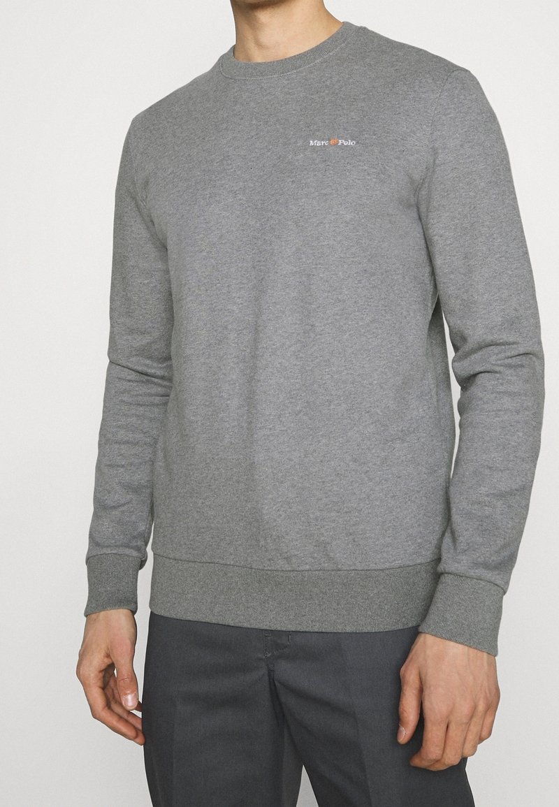 Homme portant un sweat-shirt gris uni à manches longues avec un petit logo "Marc O’Polo" sur la poitrine, associé à un pantalon gris foncé.