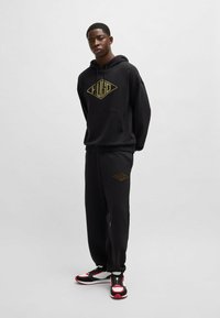 Black hoodie and sweatpants set featuring a large gold logo on the chest and thigh. Fabric is soft; fit is relaxed with an adjustable hood.