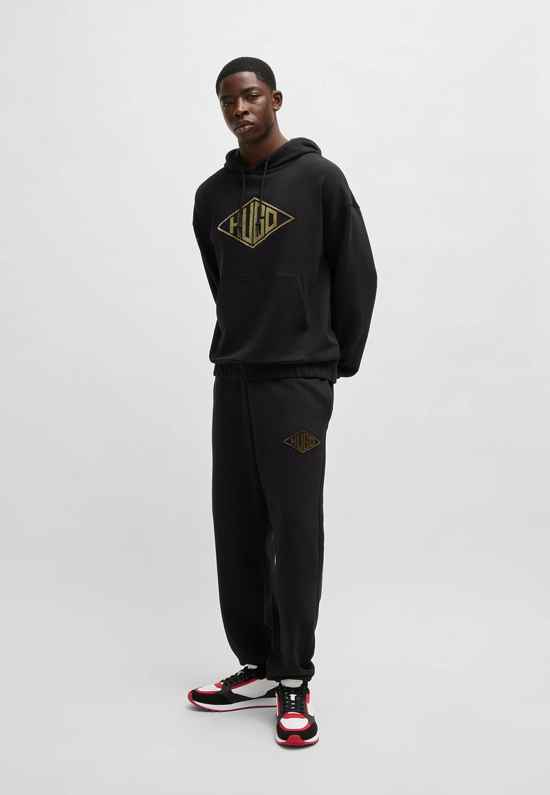 Black hoodie and sweatpants set featuring a large gold logo on the chest and thigh. Fabric is soft; fit is relaxed with an adjustable hood.