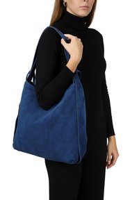 Sac en toile bleu en daim avec une texture douce, de forme rectangulaire et de larges bretelles, tenu par une personne portant un haut noir.