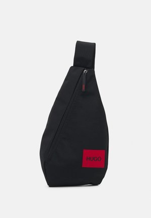 Sac en bandoulière en tissu noir de forme triangulaire. Présente un patch logo rouge et une poche zippée, avec une large sangle réglable.