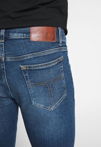 Mörkblå denimjeans med en slät textur, utrustade med en brun läderlapp på midjebandet präglad med "T/J" och standard bakfickor.