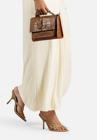Borsa a mano in pelle marrone con effetto coccodrillo, abbinata a sandali con tacco alto e cinturini color sabbia. Vestito lungo di un leggero beige.