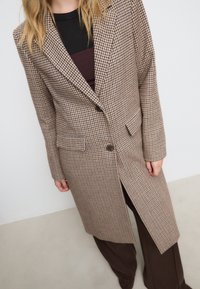 Cappotto lungo marrone a fantasia con design strutturato, caratterizzato da chiusura con un pulsante, rever e tasche laterali. Abbinato a capi scuri.