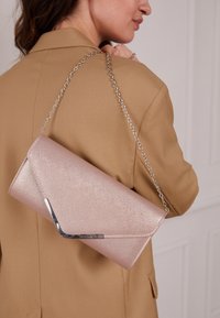 Mujer con blazer color camel sostiene un bolso de mano brillante en tonos rosa pálido con cadena plateada sobre el hombro, contra un fondo neutro.