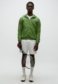 Zelena jakna z zadrgo čez belo majico, bele kratke hlače in bele nogavice; drži teniški lopar; model stoji pred enobarvnim ozadjem.