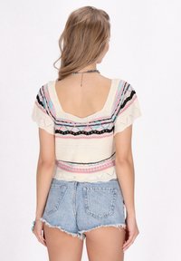 Gebreide top met korte mouwen in roomkleur met multiculturele geometrische patronen. Heeft een brede ronde rug en een gestructureerde zoom. Gecombineerd met denim shorts.