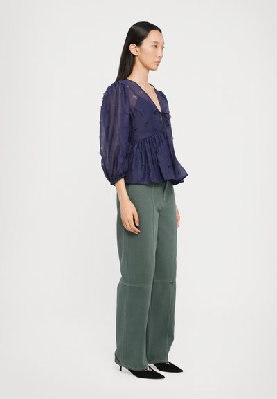Blusa navy con mangas abullonadas y dobladillo peplum, con escote en V y detalle de lazada. Combinada con pantalones de pierna ancha en color verde oliva y zapatos de tacón negros.