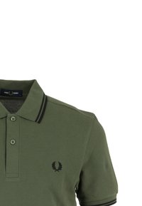 Polo en vert olive en coton avec une finition texturée, arborant un logo noir, des accents de rayure noire sur le col, et trois boutons.
