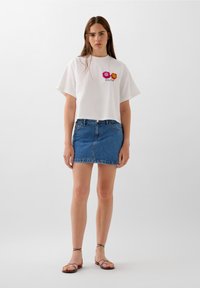 Maglietta bianca cropped con fiori ricamati, abbinata a una mini gonna di jeans blu. Il modello indossa sandali colorati con lacci.