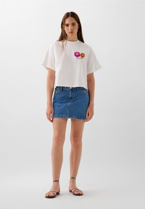 Witte cropped t-shirt met geborduurde bloemen, gecombineerd met een blauwe denim mini rok. Model draagt kleurrijke sandalen met bandjes.