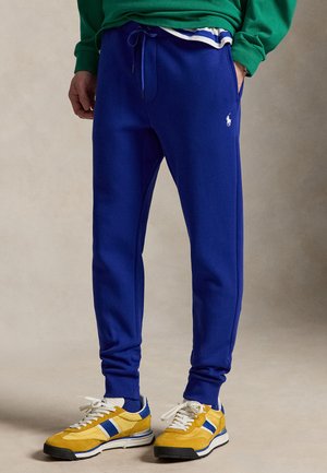 Személy kék melegítőnadrágot visel fehér logóval, sárga-kék sportcipőt és zöld pulóvert, kezét zsebében tartva áll.