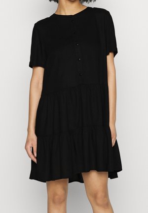 Vrouw draagt een loszittende korte zwarte jurk met knopen, korte mouwen en een gelaagde, geplooide zoom.