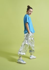 Blaues Kurzarmoberteil, glänzende silberne Cargo-Hosen mit Taschen und hellblaue Sneakers mit weißen Sohlen vor einem hellgrünen Hintergrund.