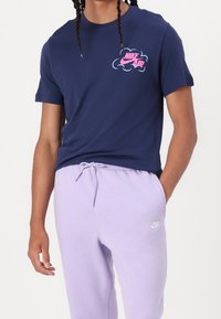 Tmavě modré bavlněné tričko se světle růžovým logem Nike a motivem mraku. Kombinováno se světle fialovými tepláky s malým bílým logem Nike.