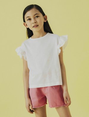 Haut blanc à manches courtes avec des accents de volants en dentelle, associé à un short rose avec un ourlet effiloché. Fond jaune vif.