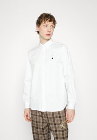 Chemise blanche à boutons avec col pointu et logo noir brodé sur la poitrine. Manches longues et coupe ajustée.