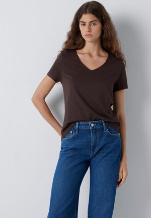 Junge Frau mit langen, welligen Haaren, die ein dunkelbraunes V-Ausschnitt-T-Shirt und blaue Jeans trägt, vor einer schlichten hellen Wand stehend.