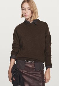 Frau trägt einen dunkelbraunen Pullover über einem schwarzen Shirt, das in einen dunkelbraunen Lederrock gesteckt ist, und hält in ihrer linken Hand eine gefaltete dunkle Tasche.