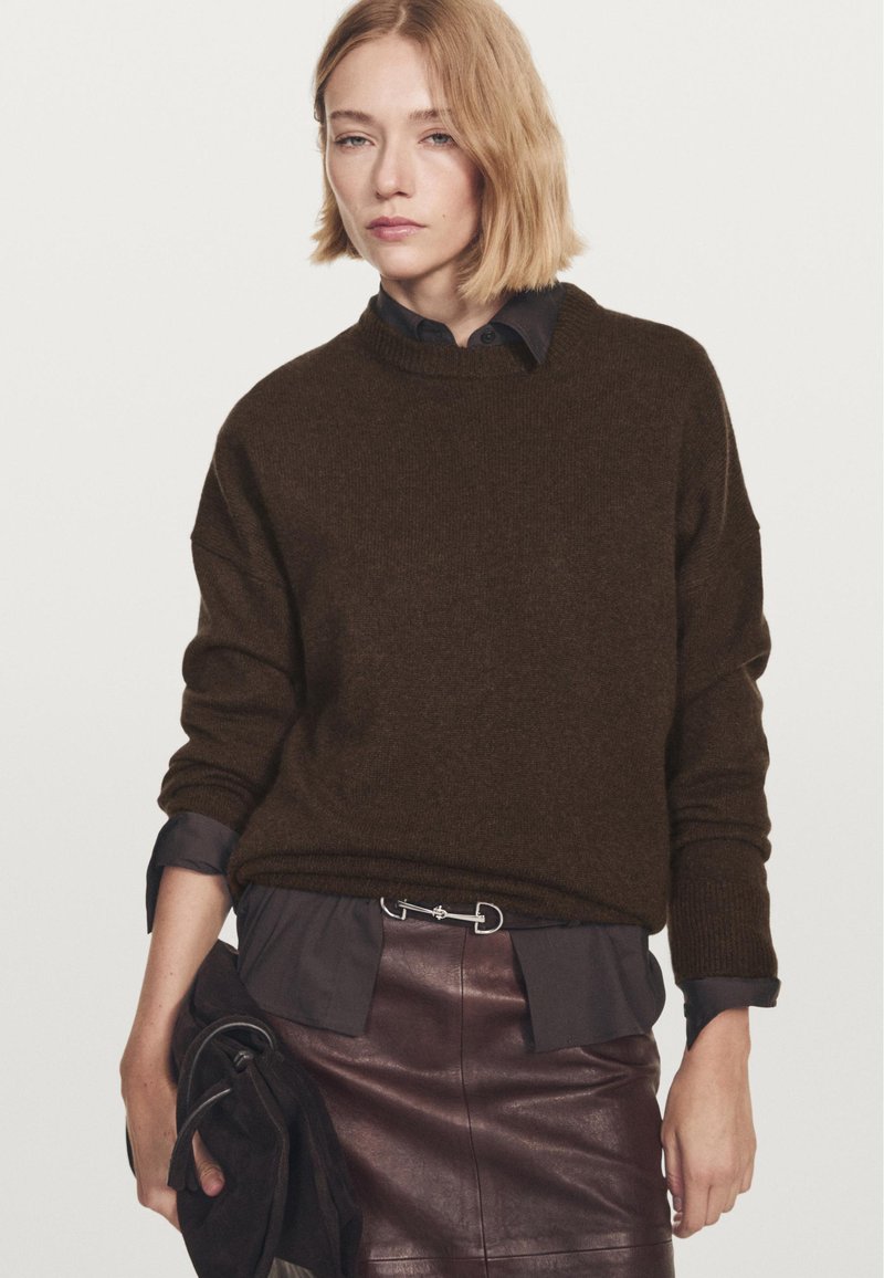 Frau trägt einen dunkelbraunen Pullover über einem schwarzen Shirt, das in einen dunkelbraunen Lederrock gesteckt ist, und hält in ihrer linken Hand eine gefaltete dunkle Tasche.