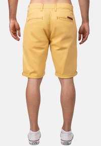Gele stoffen shorts met omgeslagen zomen, voorzien van twee achterzakken en oranje stikdetails, gecombineerd met witte sneakers.