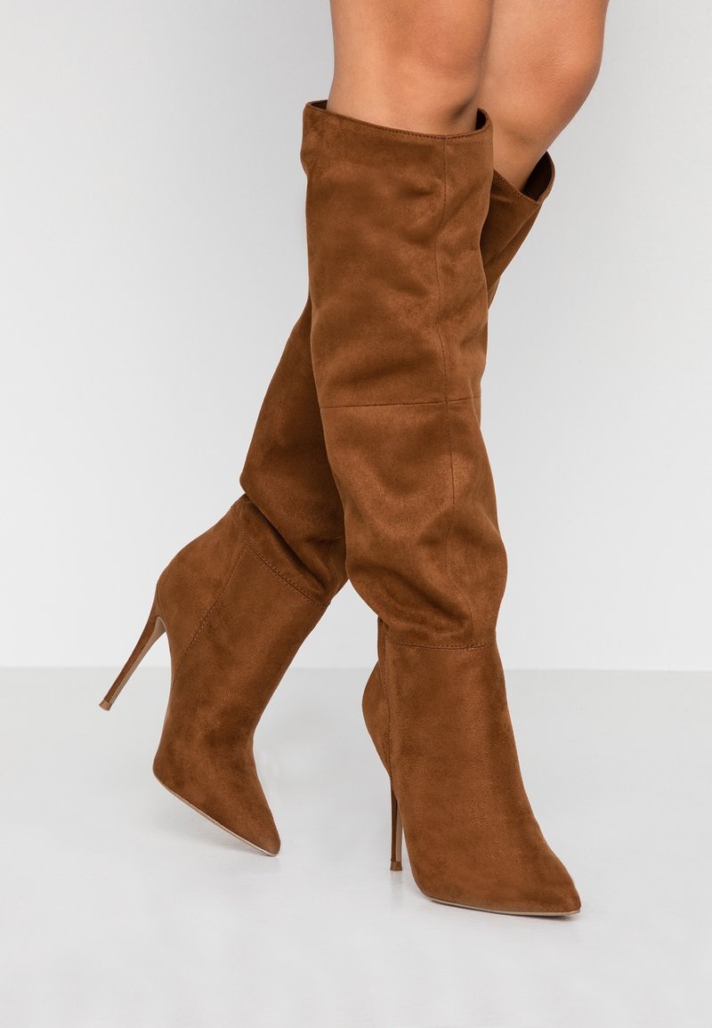 Bottes hautes en daim marron avec des bouts pointus et des talons aiguilles portées par une personne debout sur un sol blanc.