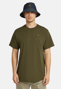 Camiseta de color verde oliva con cuello redondo y mangas cortas, combinada con un sombrero tipo bucket de color azul oscuro que presenta un parche de logo en el frente.