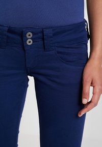 Detailní záběr na osobu v tmavě modrých kalhotách s dvojitým kovovým zapínáním a rukou odpočívající vedle stehna.